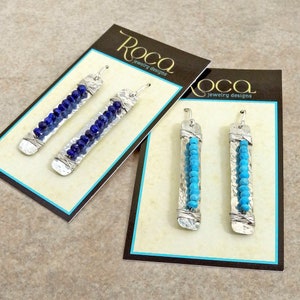 May include: Two pairs of silver dangle earrings with wire wrapped around a silver bar. One pair has blue beads and the other pair has turquoise beads. The earrings are displayed on cards with the text "Roca Jewelry Designs".