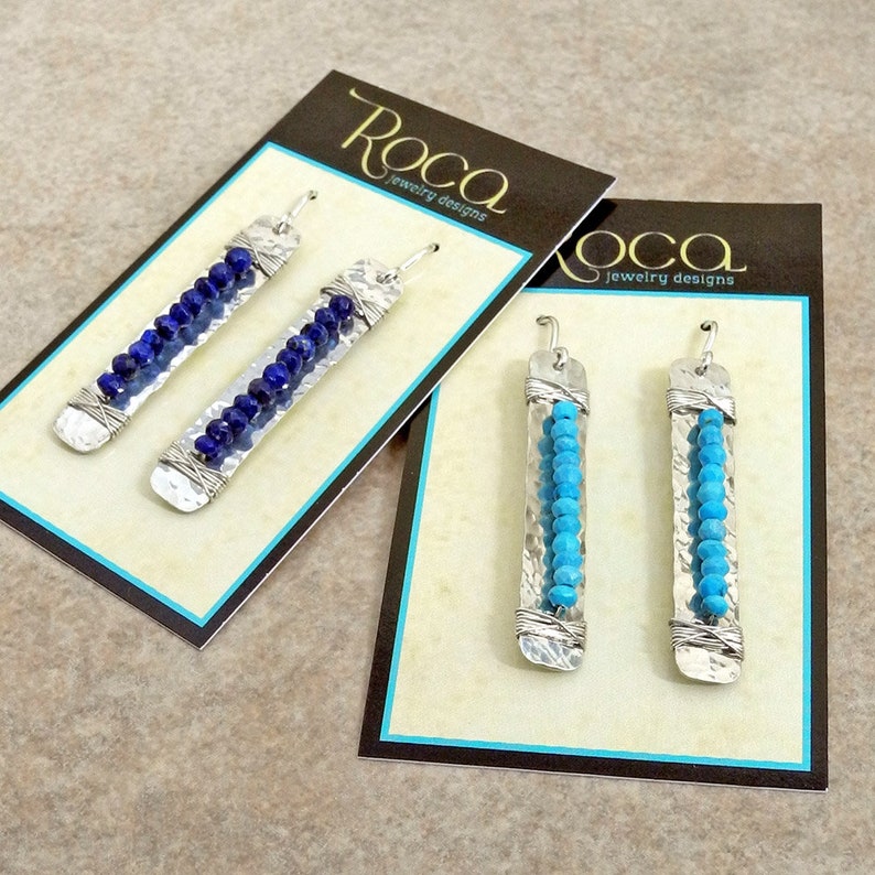 May include: Two pairs of silver dangle earrings with wire wrapped around a silver bar. One pair has blue beads and the other pair has turquoise beads. The earrings are displayed on cards with the text "Roca Jewelry Designs".