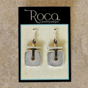 May include: Silver square earrings with a textured finish and a central hole. The earrings are displayed on a black card with the gold text "Roca jewelry designs". The earrings have silver hooks.