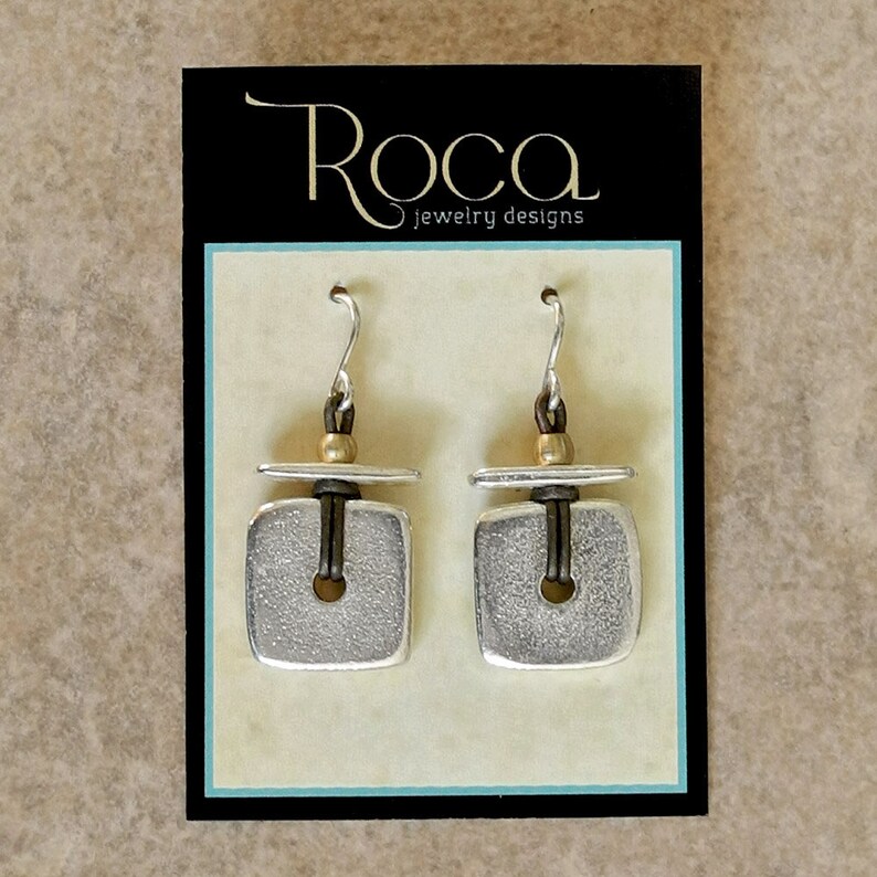 May include: Silver square earrings with a textured finish and a central hole. The earrings are displayed on a black card with the gold text "Roca jewelry designs". The earrings have silver hooks.