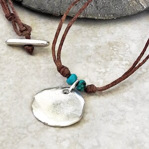May include: A silver pendant necklace with a round, organic shape and a brown leather cord. The cord has two turquoise beads.