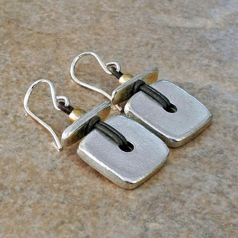 May include: Silver square earrings with a dark cord detail and a small gold bead. The earrings have silver hooks for pierced ears. The square design has a small hole in the centre. The earrings are displayed on a neutral background.