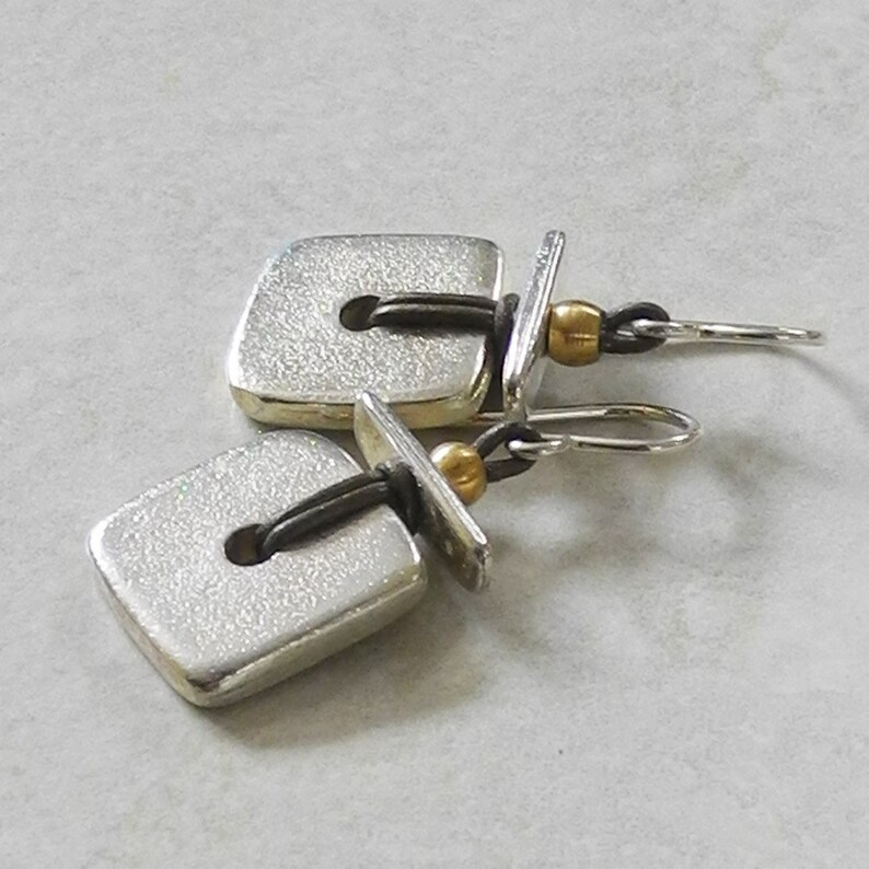 May include: A pair of silver square earrings with a textured surface. Each earring has a small, dark loop connecting to a gold bead and a silver hook. The earrings are presented on a light grey surface.