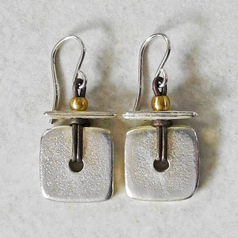 May include: A pair of silver earrings with a square, textured design. Each earring has a small, gold-coloured bead and a dark brown accent. The earrings have a hook closure.