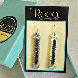 May include: A pair of silver earrings with black beads hanging from a hammered silver bar. The earrings are displayed on a card with the text "Roca Jewelry Designs".