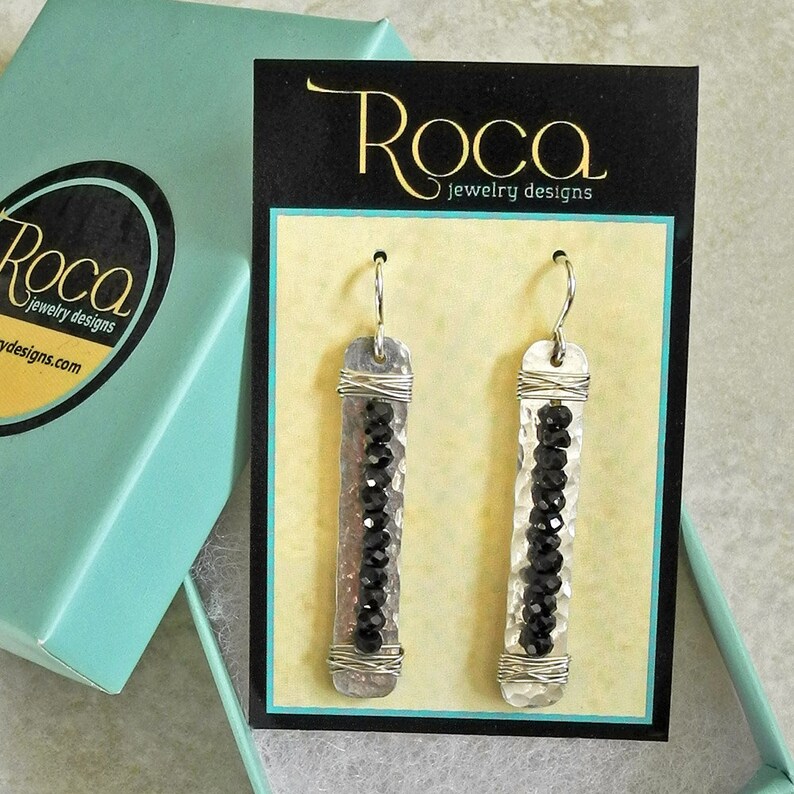 May include: A pair of silver earrings with black beads hanging from a hammered silver bar. The earrings are displayed on a card with the text "Roca Jewelry Designs".
