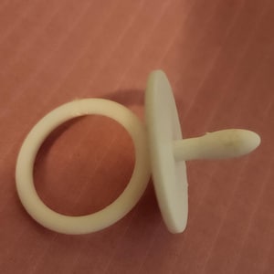May include: A white baby pacifier and teething ring are displayed against a pink background. The pacifier has a round shield and a nipple. The teething ring is a simple, circular design.