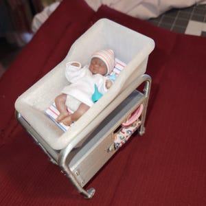 May include: A miniature baby doll in a white bassinet with a striped hat and a blue dummy. The doll is wearing a white outfit and is lying on a striped blanket. The bassinet is on a metal frame with a storage shelf.