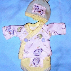 Può includere: Un set di vestiti per neonati con cappello verde chiaro, top avvolgente e pannolino. Il set è bianco con stampa di mucca e rifiniture gialle. La parte superiore ha due bottoni blu.