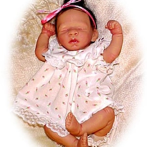 May include: A realistic baby doll, dressed in a white dress with a pink floral print and lace edging. The doll has a pink ribbon in its hair and is lying down. The doll's eyes are closed.