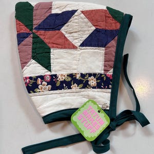 May include: A green, white, pink and blue quilted fabric bonnet with a floral print trim and a green ribbon tie. The bonnet has a tag that reads "Tatun Makes Stuff".