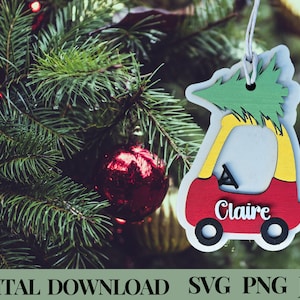 May include: Red wooden toy car Christmas ornament with a green Christmas tree on top. The car has a white interior and black wheels. The name "Claire" is written on the side of the car.
