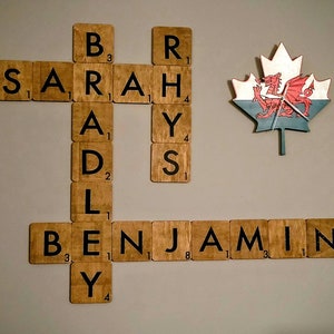 May include: A wooden wall art piece featuring the names "Sarah" and "Benjamin" spelled out using Scrabble tiles. The tiles are arranged in a cross shape with a Canadian flag in the centre.