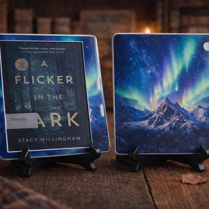 May include: Two e-readers with decorative covers. One shows the book cover of "A Flicker in the Dark" by Stacy Willingham. The other features an image of the Northern Lights over a mountain range. Both are on black stands.