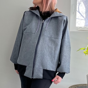 Puede incluir: Chaqueta con capucha gris con cremallera completa y ribete azul oscuro en contraste. La chaqueta tiene un panel inferior negro y un forro color canela en la capucha. Las mangas son largas y holgadas.
