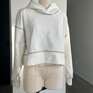 May include: White cropped hoodie with a contrasting gray stitch detail around the neckline, hood, and sleeves. The hoodie is on a dress form with the text "Royal Dress forms" printed on the front.