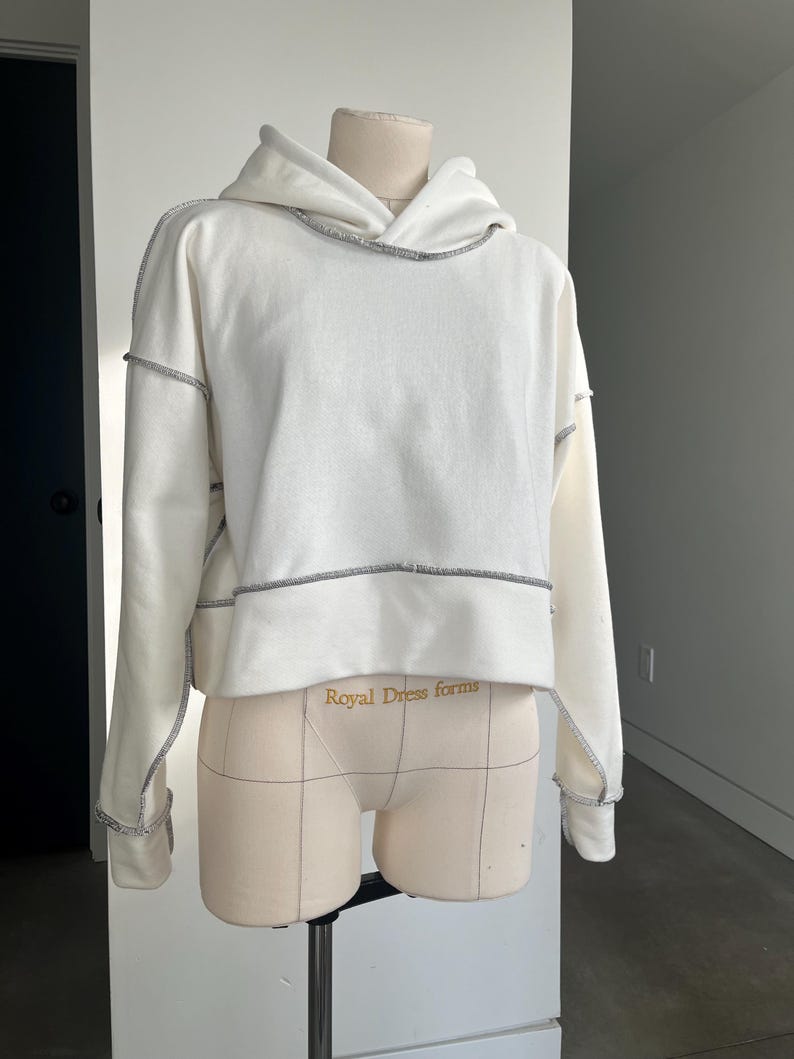 May include: White cropped hoodie with a contrasting gray stitch detail around the neckline, hood, and sleeves. The hoodie is on a dress form with the text "Royal Dress forms" printed on the front.