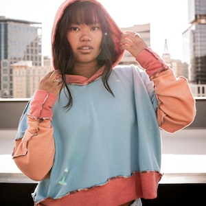May include: A woman wearing a blue, pink, and peach color block hoodie with a frayed hem. The hoodie has a drawstring hood and long sleeves.