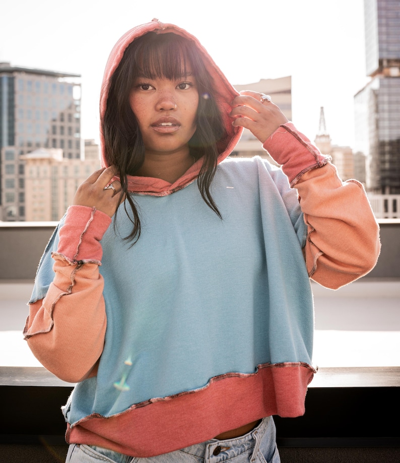 May include: A woman wearing a blue, pink, and peach color block hoodie with a frayed hem. The hoodie has a drawstring hood and long sleeves.
