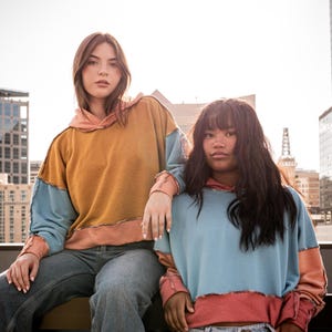 May include: Two women wearing colorful patchwork hoodies. The woman on the left is wearing a mustard yellow, light blue, and peach hoodie. The woman on the right is wearing a light blue, peach, and pink hoodie.
