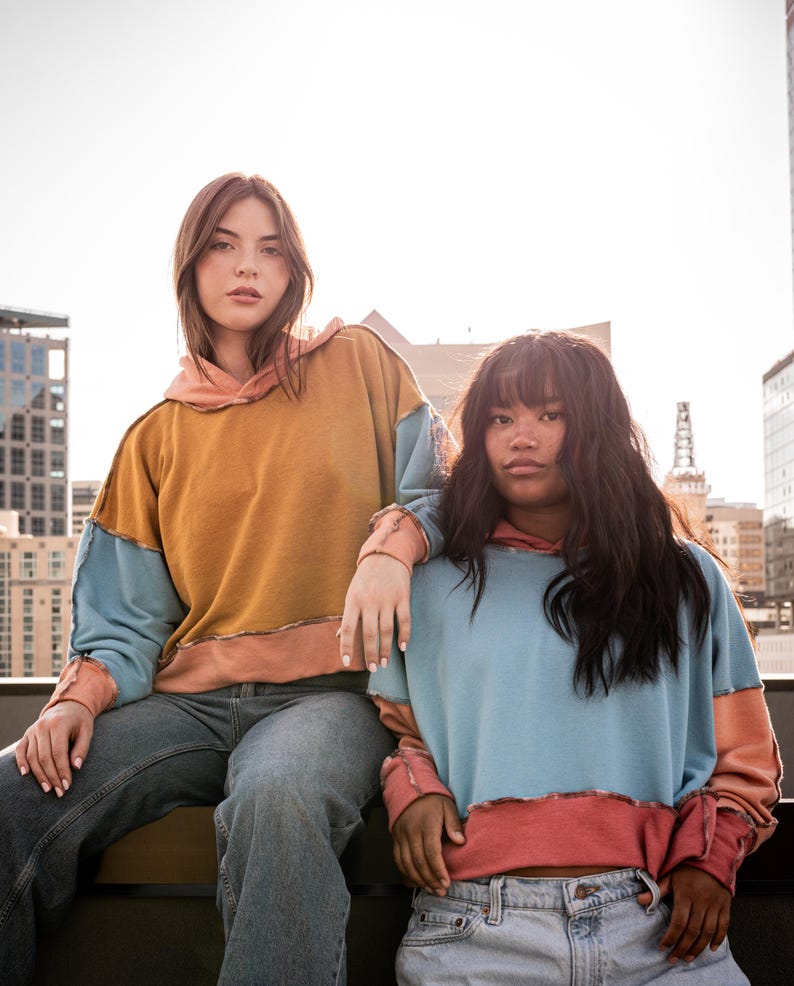 May include: Two women wearing colorful patchwork hoodies. The woman on the left is wearing a mustard yellow, light blue, and peach hoodie. The woman on the right is wearing a light blue, peach, and pink hoodie.