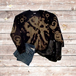 May include: A black sweatshirt with a bleached design, paired with blue denim shorts and black leather boots. The boots have a buckle detail. The items are arranged on a light brown wooden surface.