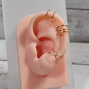 May include: Gold ear cuff jewelry with multiple rings and a criss-cross design. The jewelry is displayed on a mannequin ear.