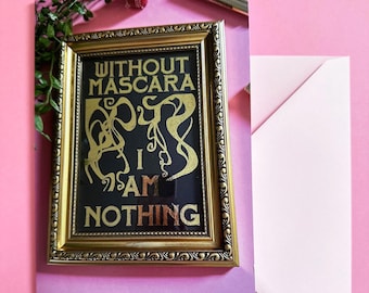 May include: A gold-framed picture with a black background and gold lettering that reads "Without Mascara I Am Nothing". The picture is on a light pink surface.
