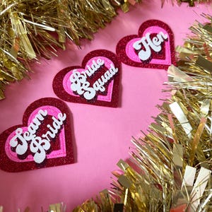 May include: Three heart-shaped decorations with glittery red and pink layers. The decorations have white text that reads "Team Bride", "Bride Squad", and "Hen". The background is pink, and gold tinsel surrounds the hearts.