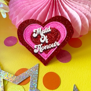 Può includere: Uno spillo a forma di cuore rosso e rosa con glitter con il testo "Maid of Honour" in bianco.
