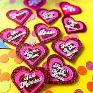 Puede incluir: Una colección de adornos de boda en forma de corazón, en tonos rosa y rojo brillante. Cada corazón presenta texto blanco, incluyendo frases como "Novia", "Novio" y "¡Recién Casados!". Confeti de varios colores rodea los corazones sobre una superficie amarilla.