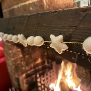 May include: A white felt garland with star and ball shapes strung on a gold cord. The garland is draped across a wooden mantel above a brick fireplace with a fire burning inside.