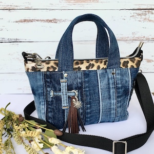 May include: A denim handbag featuring a patchwork design of various blue jean fabrics, a leopard print upper panel, two top handles, and a black adjustable shoulder strap. A brown leather tassel with a silver sun charm hangs from the front.