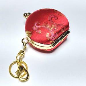 Könnte beinhalten: Eine rote, runde Geldbörse mit goldenen Akzenten und einem goldenen Schlüsselanhänger. Die Geldbörse hat ein verziertes Blumenmuster in Gold und Rosa. Der Verschluss ist gold und schwarz. Der Schlüsselanhänger hat einen Karabinerverschluss und einen Schlüsselring.