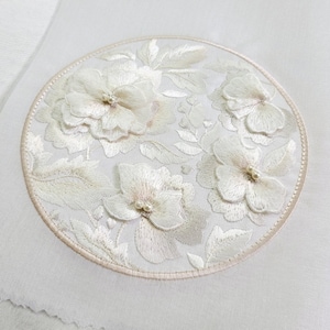 Peut inclure: Un tissu blanc brodé avec un motif circulaire de fleurs et de feuilles. Les fleurs sont en relief et ont des accents nacrés.
