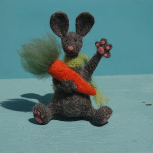May include: A handmade felt mouse figurine in gray, holding a large orange carrot with green leafy top. The mouse wears a green scarf and has pink paw pads. The figurine is set against a light blue background.
