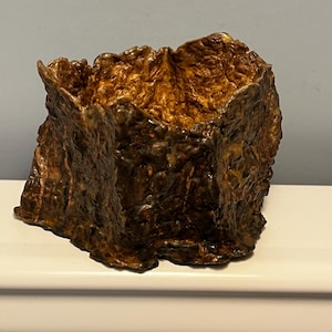 May include: A textured, square-shaped decorative bowl with a rough, organic appearance. The bowl is primarily brown with bronze and gold highlights, creating a warm, earthy aesthetic. The interior is a lighter shade of gold.