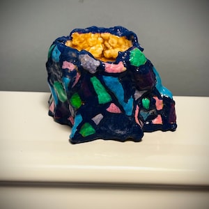 May include: A small, abstract ceramic vessel with a textured, mosaic-like exterior. The vessel is primarily dark blue, with colorful patches of green, pink, and purple. The interior is a warm, golden color.