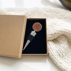 May include: A silver-toned metal bottle stopper with a circular brown leather top featuring the engraved letters S and J with the number 20. The stopper is presented inside a rectangular brown cardboard gift box with a black velvet insert.