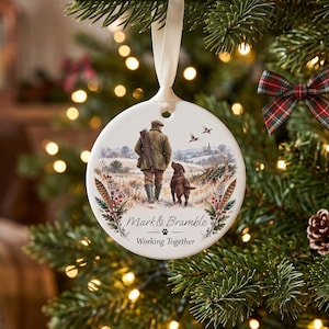 May include: A round white ceramic Christmas ornament hanging by a cream ribbon. The ornament features a printed illustration of a person in a green jacket walking with a brown dog in a field, with the text Mark & Bramble and Working Together.