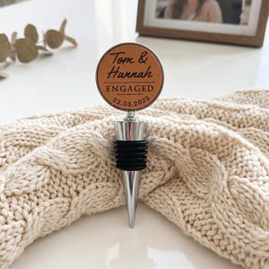 May include: A circular bottle stopper featuring a tan leather-like insert with the engraved text Tom & Hannah ENGAGED 22.03.2022. The stopper has a silver-toned metal top and a black ribbed silicone base for sealing bottles securely.