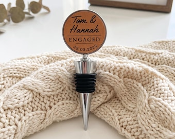 May include: A circular bottle stopper featuring a tan leather-like insert with the engraved text Tom & Hannah ENGAGED 22.03.2022. The stopper has a silver-toned metal top and a black ribbed silicone base for sealing bottles securely.