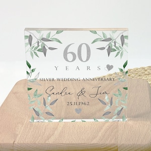 May include: A clear acrylic block with a printed design celebrating a 60th wedding anniversary. The design includes the text "60 YEARS SILVER WEDDING ANNIVERSARY" with the names "Sandra & Jim" and the date "25.11.1962". The block is set on a wooden base.