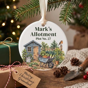 May include: A round white ceramic ornament hanging from a cream ribbon, featuring a printed illustration of a garden shed, a wheelbarrow filled with vegetables, and a robin. The text reads Mark's Allotment Plot No. 27 in black lettering.