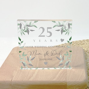 May include: A clear acrylic block commemorating a 25th wedding anniversary. The block features the text "25 YEARS SILVER WEDDING ANNIVERSARY" with the names "Mum & Dad" and the date "21.09.1999". The design includes green and gray leaf accents.