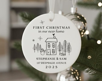 May include: A white ceramic Christmas ornament with a ribbon, hanging on a Christmas tree. The ornament has black text and a house illustration that reads "FIRST CHRISTMAS in our new home" with a name and address.
