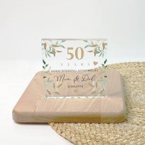 May include: A clear acrylic block with gold text and leaf accents, celebrating a 50th wedding anniversary. The block reads "50 YEARS GOLD WEDDING ANNIVERSARY" with the names "Mum & Del" and the date "11.06.1974". It rests on a light wooden base.