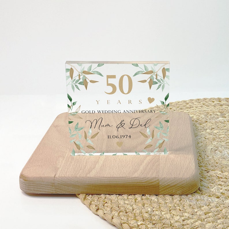 May include: A clear acrylic block with gold text and leaf accents, celebrating a 50th wedding anniversary. The block reads "50 YEARS GOLD WEDDING ANNIVERSARY" with the names "Mum & Del" and the date "11.06.1974". It rests on a light wooden base.