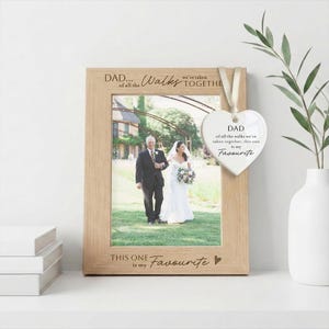 May include: A light wood picture frame with the engraved text DAD... of all the WALKS we've taken TOGETHER THIS ONE is my Favourite. A white heart-shaped ceramic ornament hangs from the frame with the same text printed in black ink.