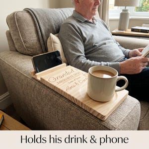 May include: A light-colored wooden armrest tray featuring a slot for a smartphone and a circular indentation for a mug. The surface is engraved with the text "Grandad's FAVOURITE SPOT lots of love Alma & Dexter" and includes a small graphic icon.